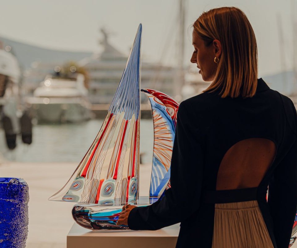 A woman at an art exhibition in Porto Montenegro, thoughtfully examining a colorful glass sculpture shaped like a sailboat, with luxurious yachts and the marina backdrop enhancing the artistic ambiance.