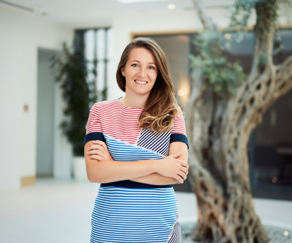 A woman in a striped dress smiles confidently with her arms crossed in a modern indoor space featuring a prominent tree.