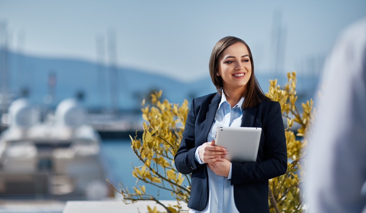 A professional woman in a navy blazer holds a tablet and smiles, with a marina in the background.