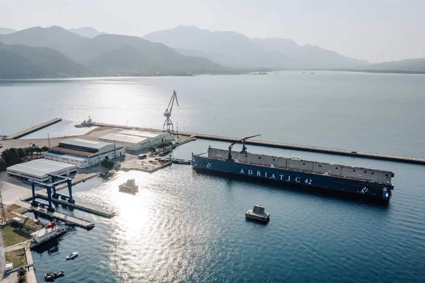 Aerial view of the Adriatic 42 shipyard, showcasing the shipyard’s facilities, a large floating dock labeled "Adriatic 42," and the surrounding scenic bay and mountains.
