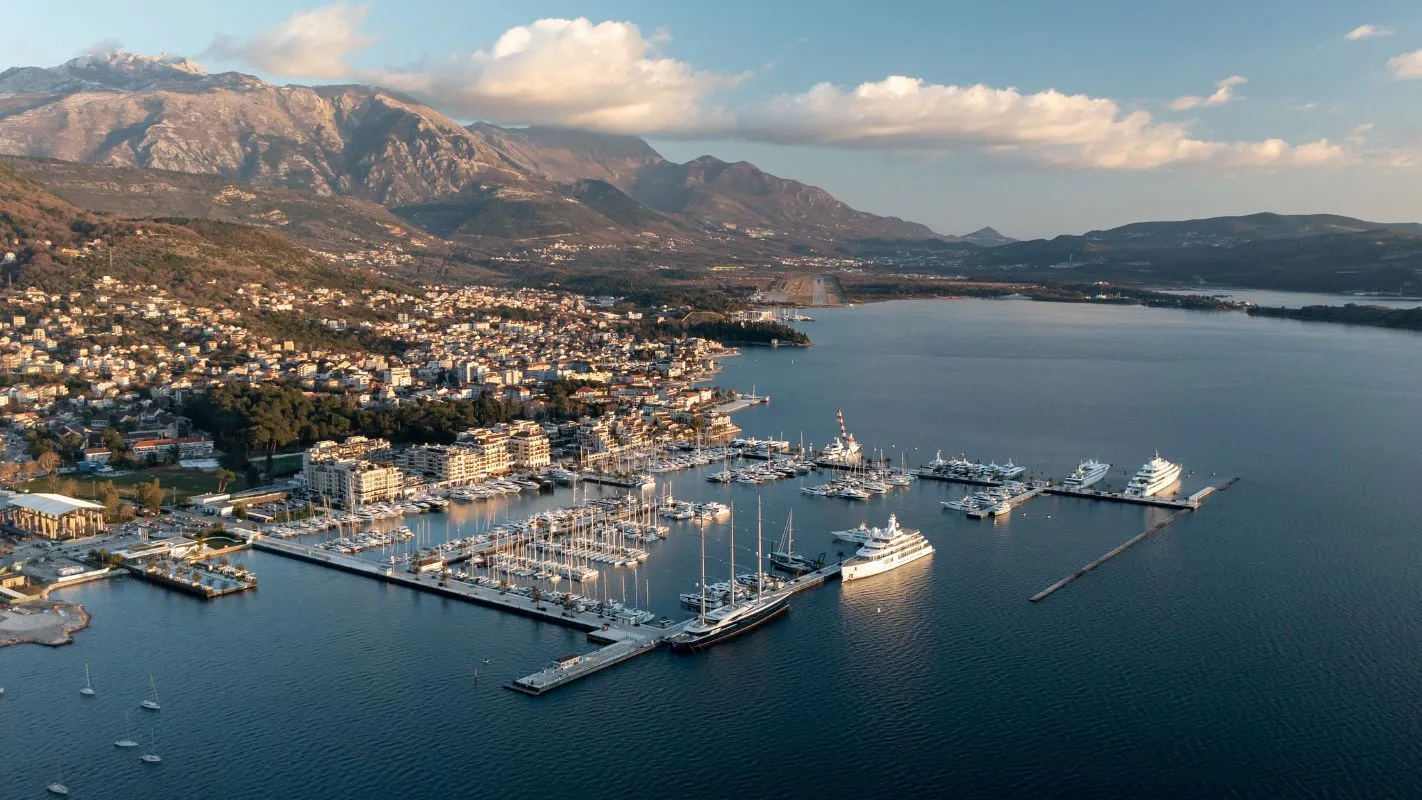 Aerial view of a luxurious marina nestled between a coastal town and majestic mountains, showcasing an array of yachts and sailboats.