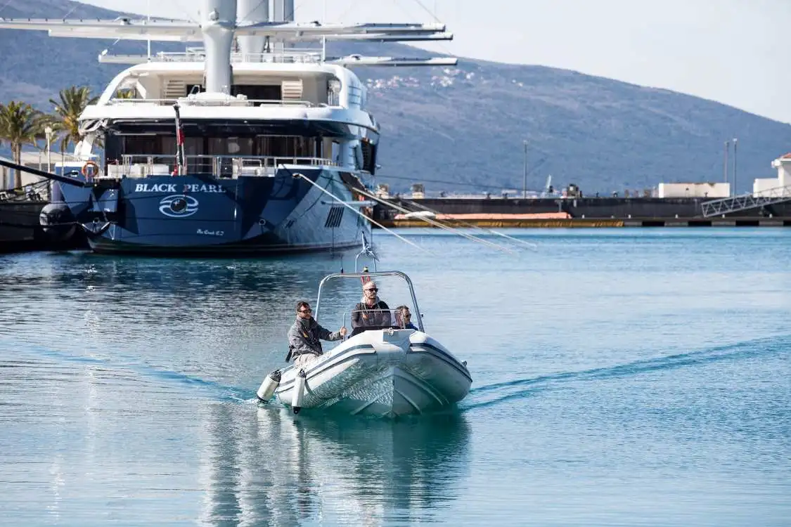 A small grey tender boat with three people aboard is moving through the calm marina waters, leaving a gentle wake behind. In the background, a large luxury sailing yacht named "Black Pearl" is docked, with its sleek dark hull and multiple masts. Hills and a marina pier are visible in the distance.