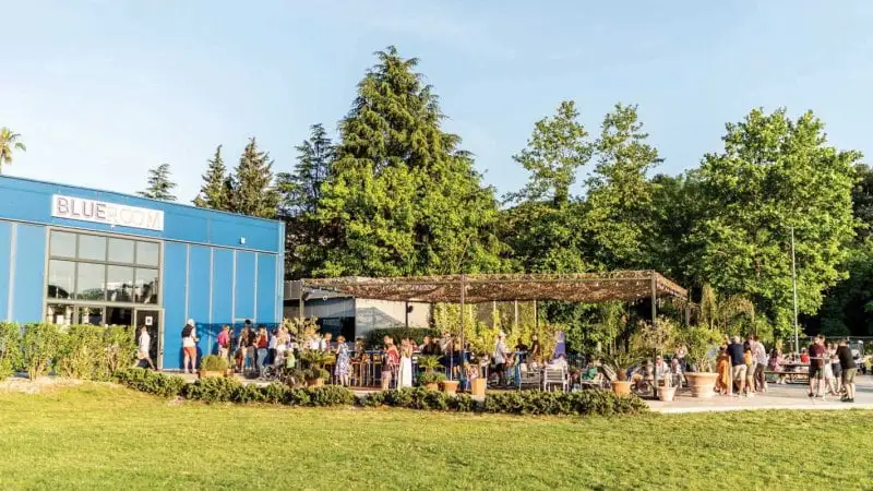 A vibrant outdoor cafe with people socializing under a pergola beside a modern blue building, surrounded by lush greenery on a sunny day.