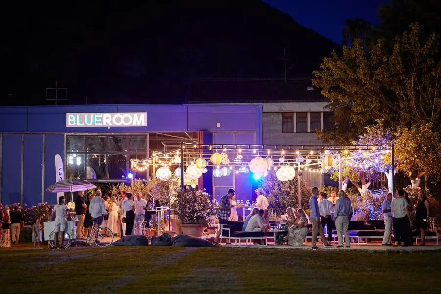 Nighttime outdoor gathering at the Blue Room venue, with guests mingling under string lights and colorful lanterns.