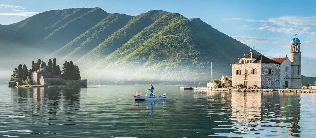 A peaceful morning scene of a mountain-backed bay with a man rowing a boat between two small, historic island churches surrounded by calm waters.