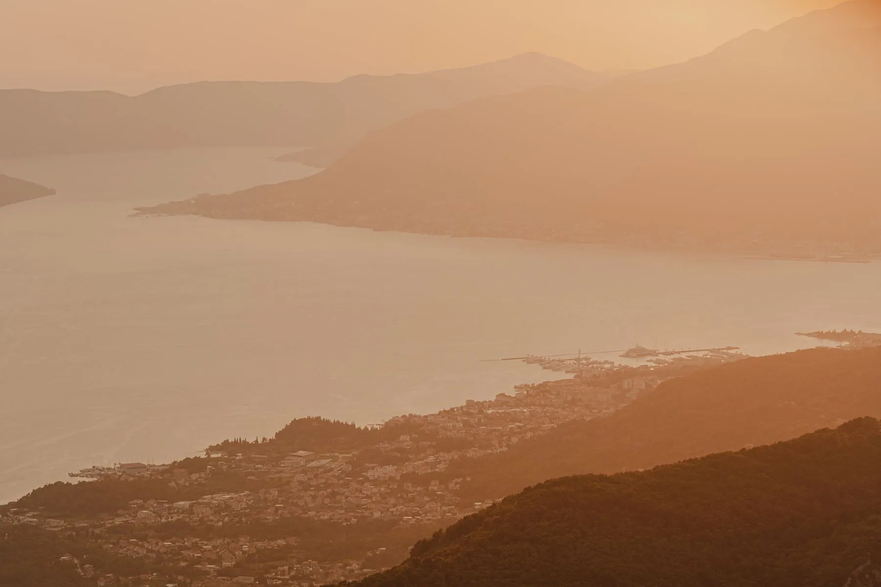 Serene aerial view of Boka Bay at sunset, with warm golden light casting a soft glow over the tranquil waters and surrounding mountains, creating a peaceful, ethereal landscape.