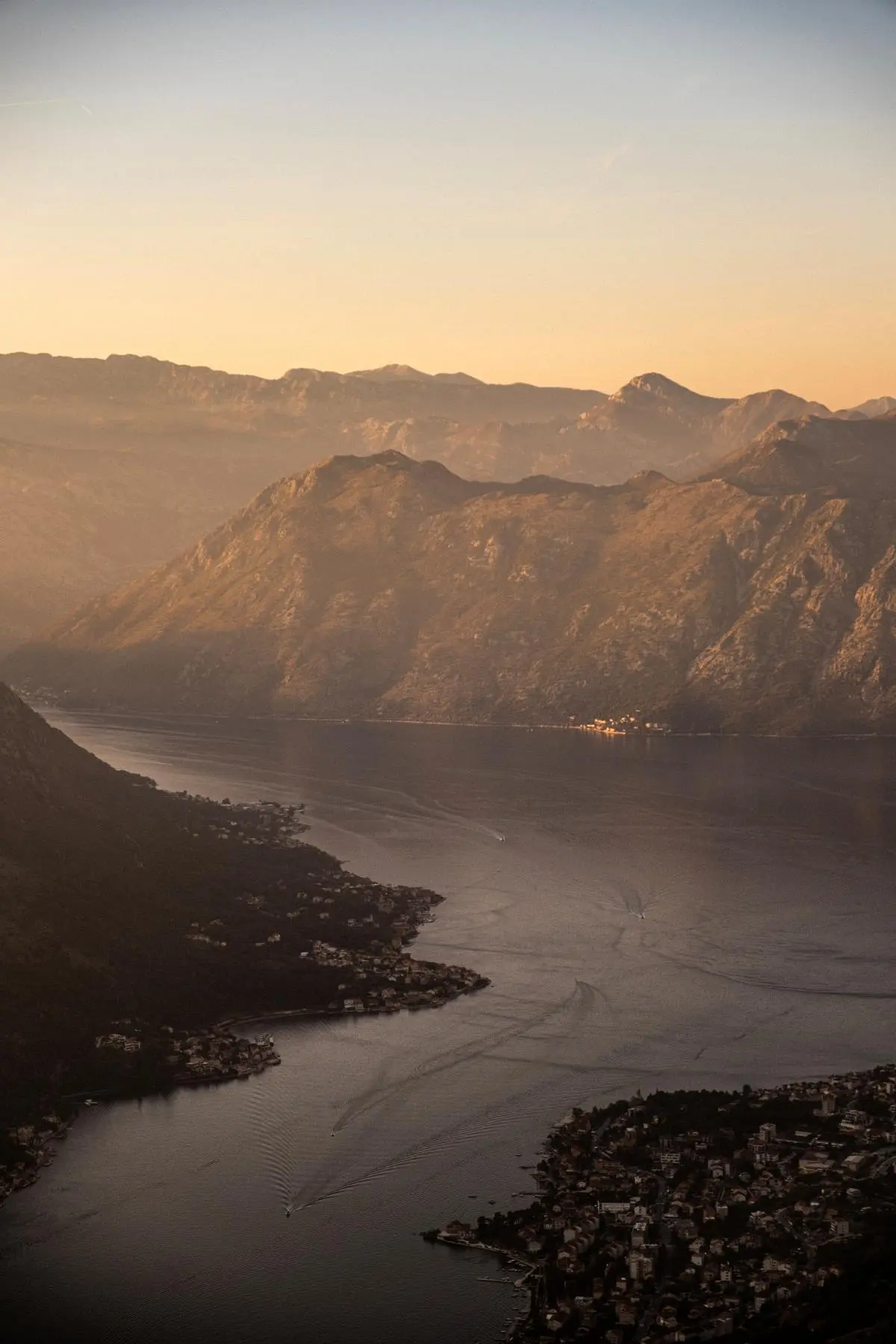 A breathtaking aerial view of a serene bay surrounded by majestic mountain ranges, captured during a golden sunset, with small coastal settlements dotting the shoreline.