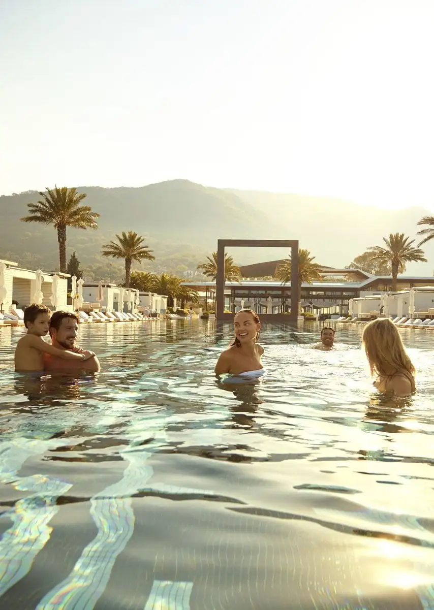 People enjoying a swim in a luxurious outdoor pool at sunset, surrounded by palm trees and cabanas, with mountains in the background, creating a serene and relaxing atmosphere.