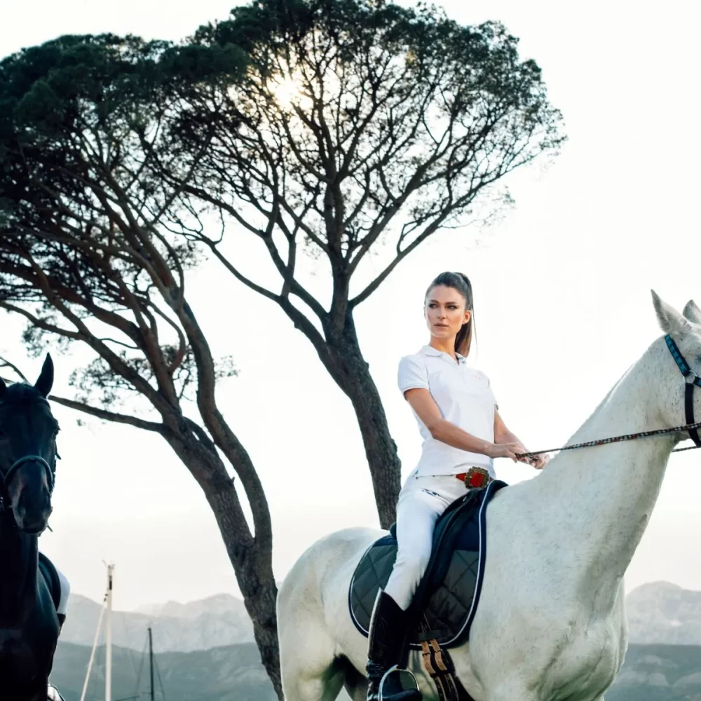 A couple horseback riding with a man on a black horse and a woman on a white horse, under a tree with mountains in the background.