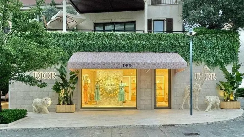 Exterior of a Dior store with a stone facade, decorated with lush greenery, a patterned awning, and statues of a lion and a sun emblem by the entrance.