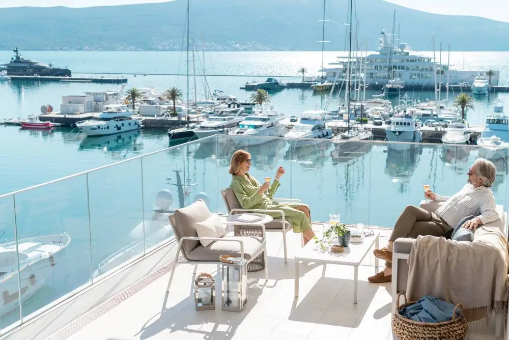 Couple sits on a luxurious balcony overlooking a Porto Montenegro marina filled with yachts, enjoying drinks and a peaceful conversation. The scene captures a serene moment of relaxation with a backdrop of calm waters and distant mountains under clear skies.