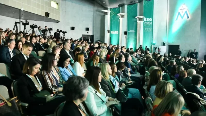 A large audience attends the ESG Adria Summit Montenegro 2024, seated in a conference hall with green-lit branding. Attendees in business attire listen attentively, while cameras capture the event.