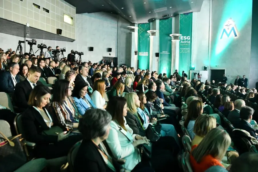 A large audience attends the ESG Adria Summit Montenegro 2024, seated in a conference hall with green-lit branding. Attendees in business attire listen attentively, while cameras capture the event.