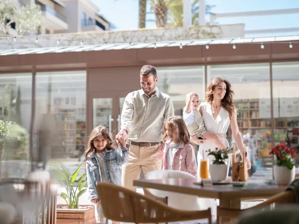 A smiling family of four entering restaurant with the father holding one daughter's hand and the mother carrying their young child. The scene is filled with warmth and happiness, capturing a moment of togetherness in a stylish, contemporary setting.