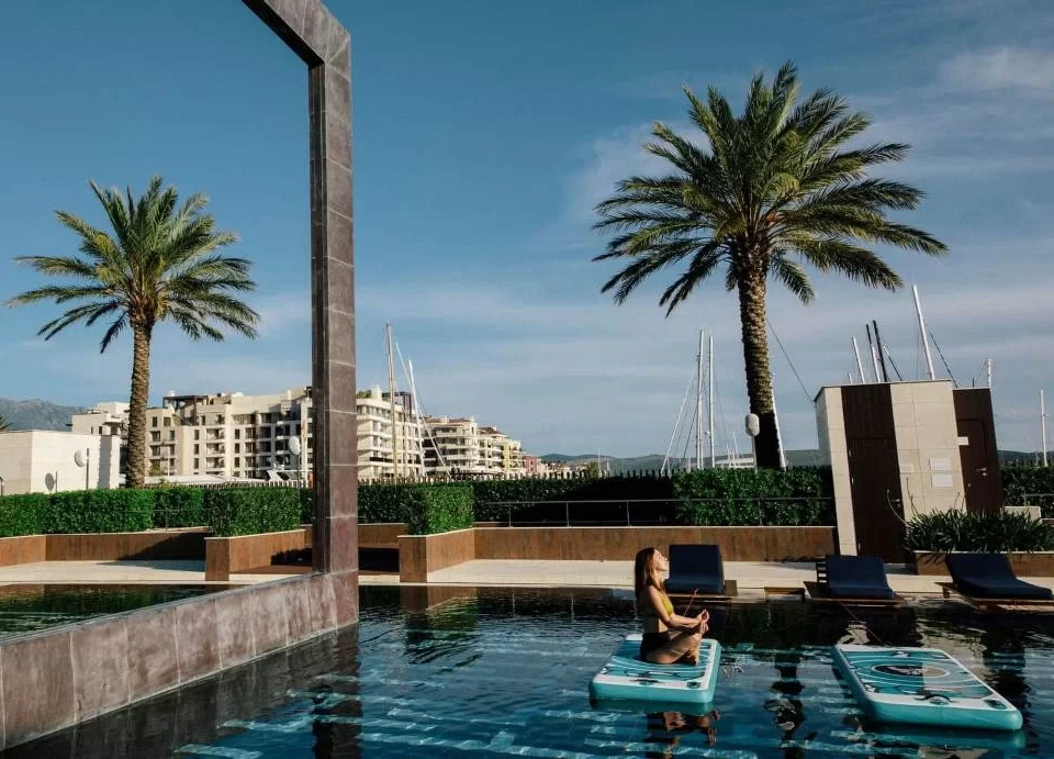 A woman practicing floating yoga on a paddleboard in the elegant infinity pool at Porto Montenegro, surrounded by palm trees, luxury residences, and marina views under a clear blue sky.