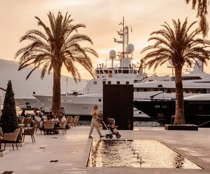 A tranquil sunset at a marina with yachts docked near palm trees. A woman pushes a stroller past a water feature, while diners enjoy the outdoor cafe.