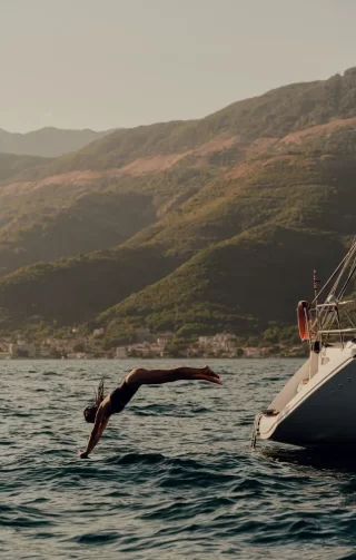 A woman gracefully dives from a sleek yacht into the calm, blue waters, set against a backdrop of verdant mountains. The scene exudes a sense of freedom and adventure, capturing the luxury of a spontaneous escape in an exclusive, natural paradise.