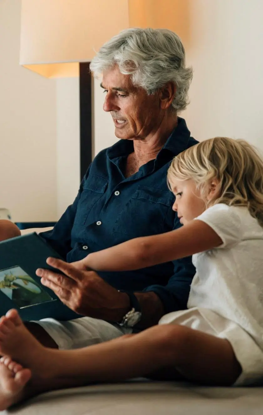 An older man with gray hair reads a book to his young grandchild, who is sitting close beside him, both deeply engaged in the story. The cozy indoor setting is softly lit by a lamp, creating a warm, intimate moment.