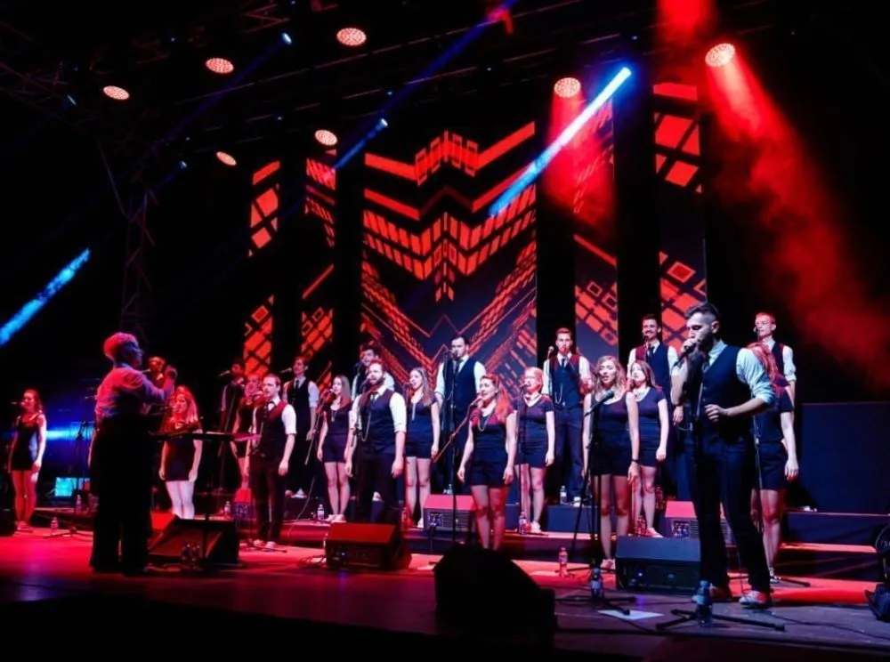 Choir performing on stage under vibrant red and blue lighting, with a conductor leading and singers dressed in formal black attire.