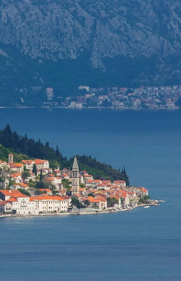 A scenic view of the historic town of Perast in the Bay of Kotor, Montenegro, featuring charming stone houses with red-tiled roofs, a prominent church bell tower, and a mountainous backdrop.