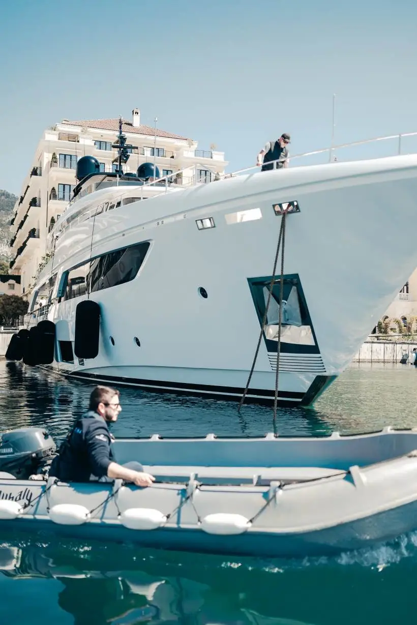 A luxury white yacht docked at a marina with a modern waterfront building in the background. A crew member is standing on the yacht's deck, adjusting ropes, while another man in a small grey motorized dinghy moves through the water in the foreground. The scene is illuminated by bright sunlight, reflecting off the calm water.