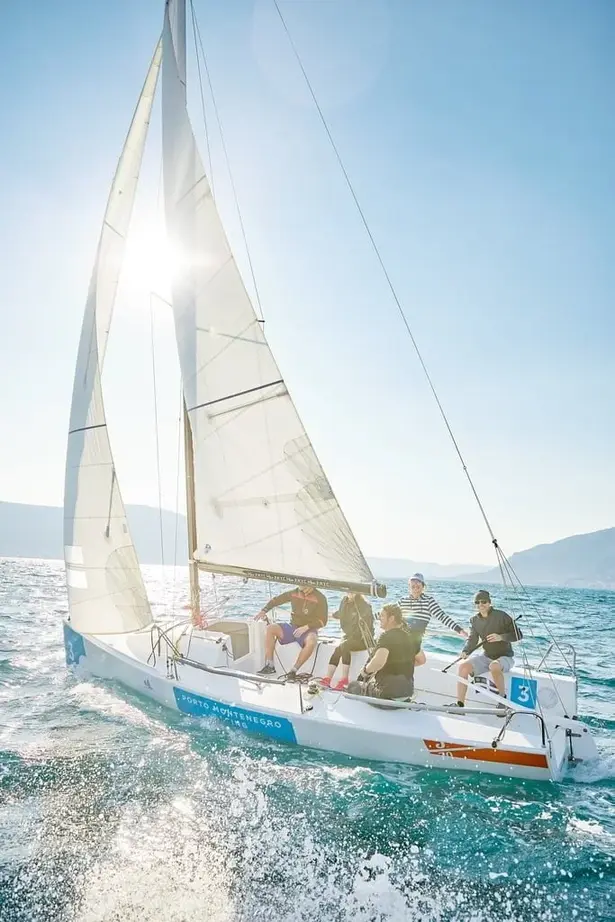 A group of sailors enjoying a thrilling sailing experience on a Porto Montenegro-branded sailboat, with the sun shining through the sails and waves splashing against the hull.