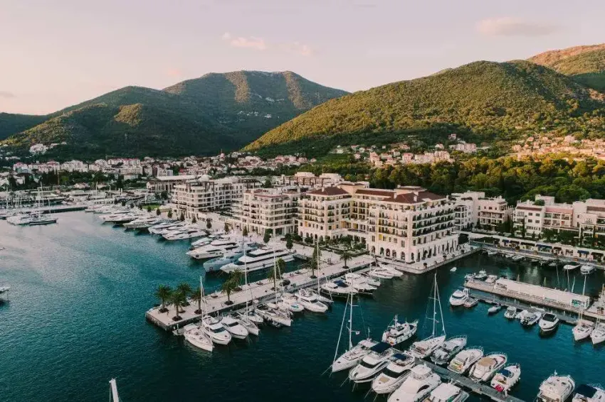Aerial view of a luxury marina with yachts, Mediterranean-style buildings, and lush green hills at sunset.