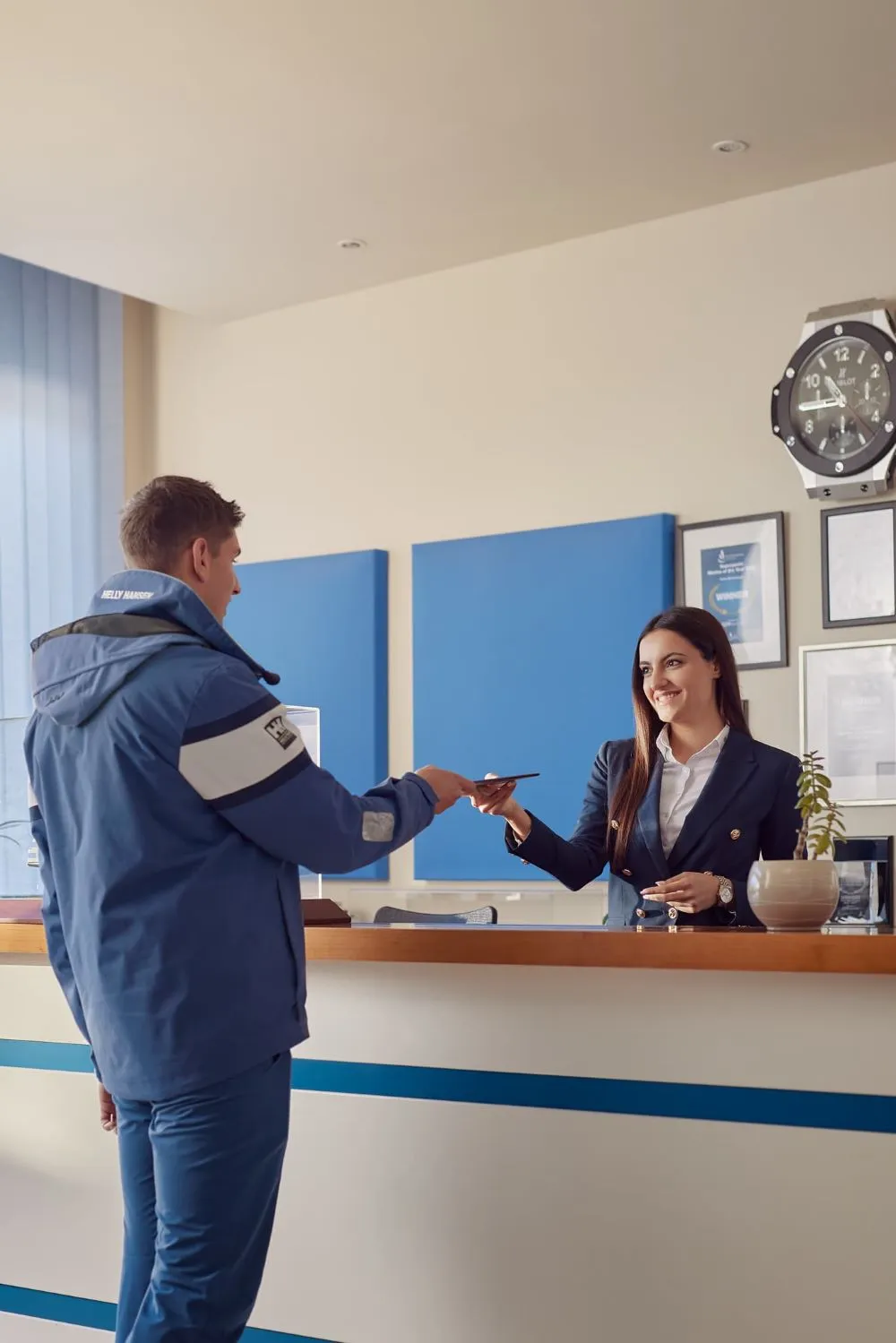 A marina office reception where a male visitor in a blue Helly Hansen jacket is handing over his passport or documents to a smiling female receptionist in a navy blue blazer. The modern interior has a nautical theme with blue panel decorations, a large wall clock, and framed certificates.