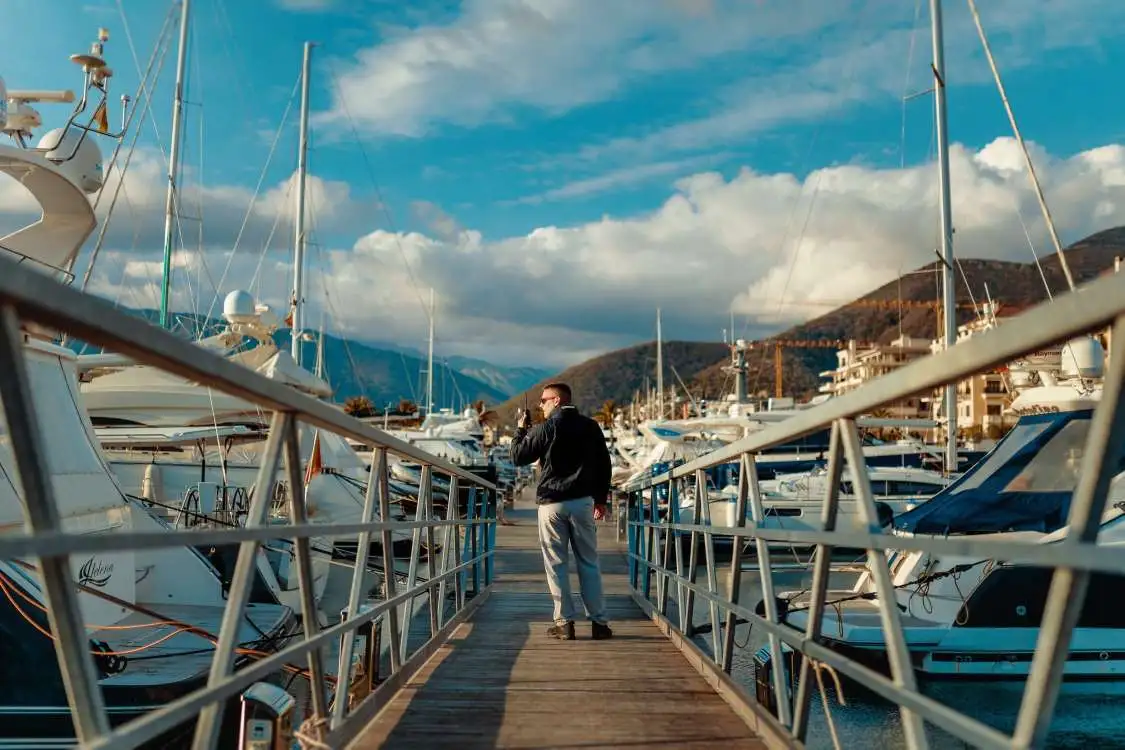 A marina security officer in a black jacket and grey pants walks along a wooden dock surrounded by luxury yachts. He is speaking into a walkie-talkie while the backdrop features mountains, waterfront buildings, and a partly cloudy sky.