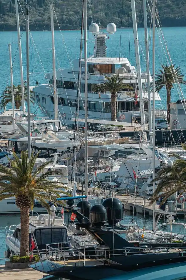 A marina filled with luxury yachts, with a large white superyacht prominently visible in the background. Palm trees line the dock, and multiple sailboats with tall masts are moored in the harbor. The turquoise sea and lush green hills in the background enhance the scenic setting.