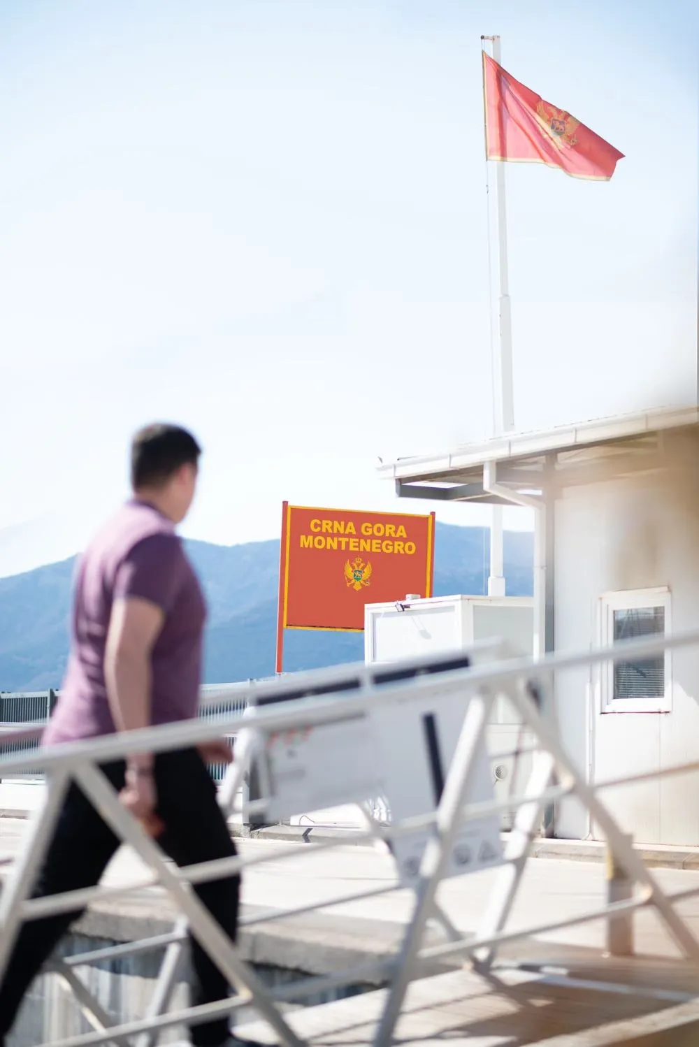 A person walking on a gangway towards a port entry in Montenegro. A red sign with the words "CRNA GORA MONTENEGRO" and the national coat of arms is visible, along with a flag of Montenegro waving on a pole. Mountains form a backdrop to the scene.