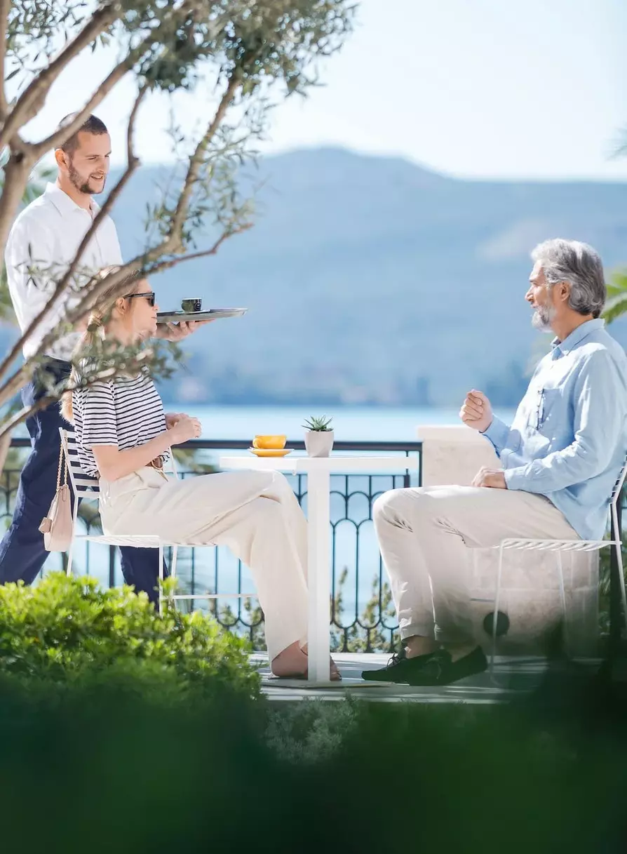 Couple enjoying morning coffee at an outdoor seaside café, served by a waiter, with a backdrop of mountains and water, surrounded by lush greenery.