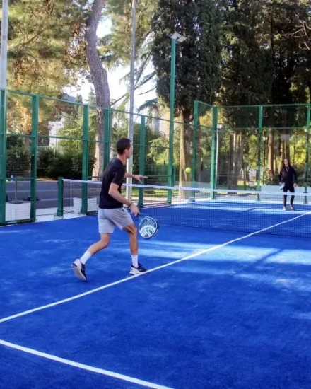 A young man in black sportswear plays padel on a vibrant blue outdoor court, hitting the ball while his opponent, a woman dressed in black, waits on the other side of the net, surrounded by lush greenery.