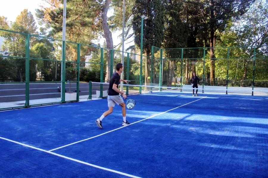 A young man in black sportswear plays padel on a vibrant blue outdoor court, hitting the ball while his opponent, a woman dressed in black, waits on the other side of the net, surrounded by lush greenery.