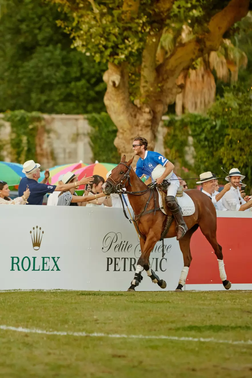Polo player on horseback greeting spectators by the field during a match, with a Rolex advertisement in the background and colorful umbrellas providing shade.