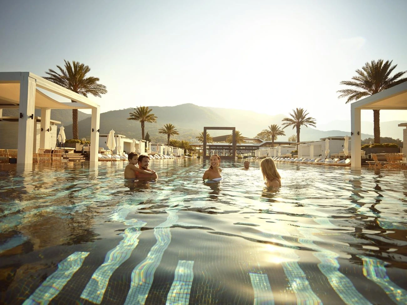 A group of people enjoying a relaxing swim in the luxurious infinity pool at Porto Montenegro, surrounded by elegant cabanas, palm trees, and a stunning mountain backdrop during golden hour