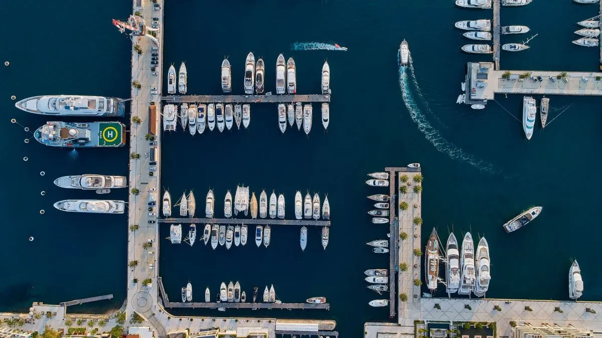 A top-down aerial view of Porto Montenegro Marina, featuring neatly arranged yachts and superyachts docked along the jetties, with boats navigating through the deep blue waters.
