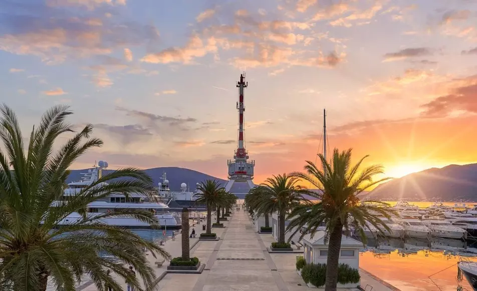 Sunset view of a marina jetty lined with palm trees, luxury yachts docked on either side, and a large crane structure at the end, with mountains in the background and a colorful sky.