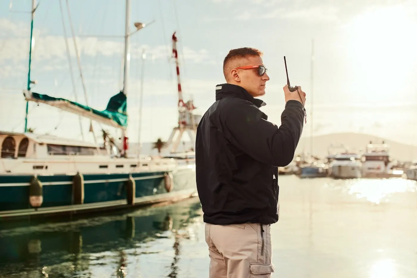 A marina security officer or dockmaster wearing a black jacket and sunglasses holds a walkie-talkie while overseeing the Porto Montenegro marina. Behind him, luxury yachts and sailboats are moored, reflecting the golden sunlight on the calm water, with maritime cranes and distant hills visible in the background.