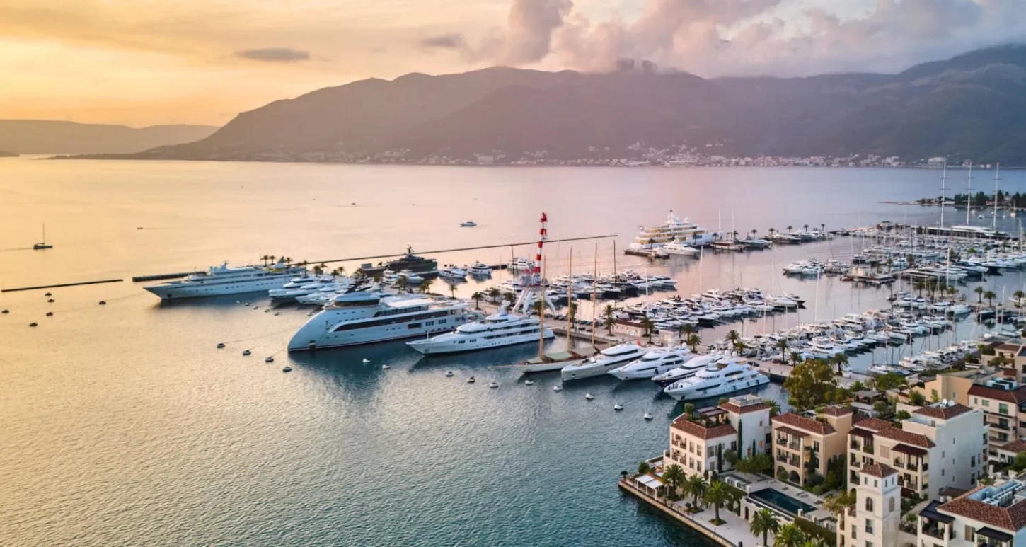 Aerial view of a luxury marina at sunset, featuring rows of large yachts and boats docked near a waterfront lined with upscale buildings. The calm sea reflects the soft orange and pink hues of the sky, with mountains in the background and palm trees scattered along the shore.