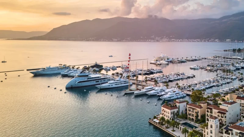 Aerial view of a luxury marina at sunset, featuring rows of large yachts and boats docked near a waterfront lined with upscale buildings. The calm sea reflects the soft orange and pink hues of the sky, with mountains in the background and palm trees scattered along the shore.