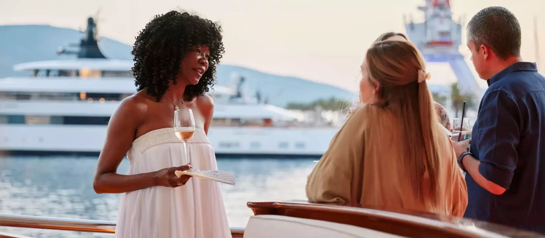 Three elegantly dressed individuals engage in a relaxed conversation on a luxury yacht's deck, with one woman in a white dress holding a glass of wine. The serene Porto Montenegro marina in the background features a sleek superyacht, underscoring an atmosphere of exclusivity and sophistication, set against the soft hues of a sunset.