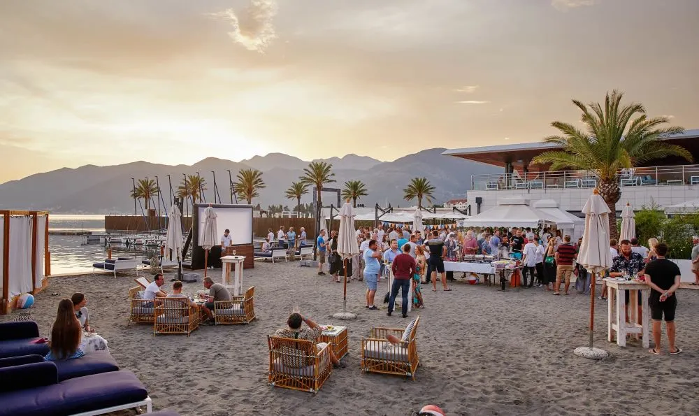 A stylish beach event during sunset, with guests enjoying an elegant outdoor gathering surrounded by palm trees, waterfront views, and scenic mountains in the background.