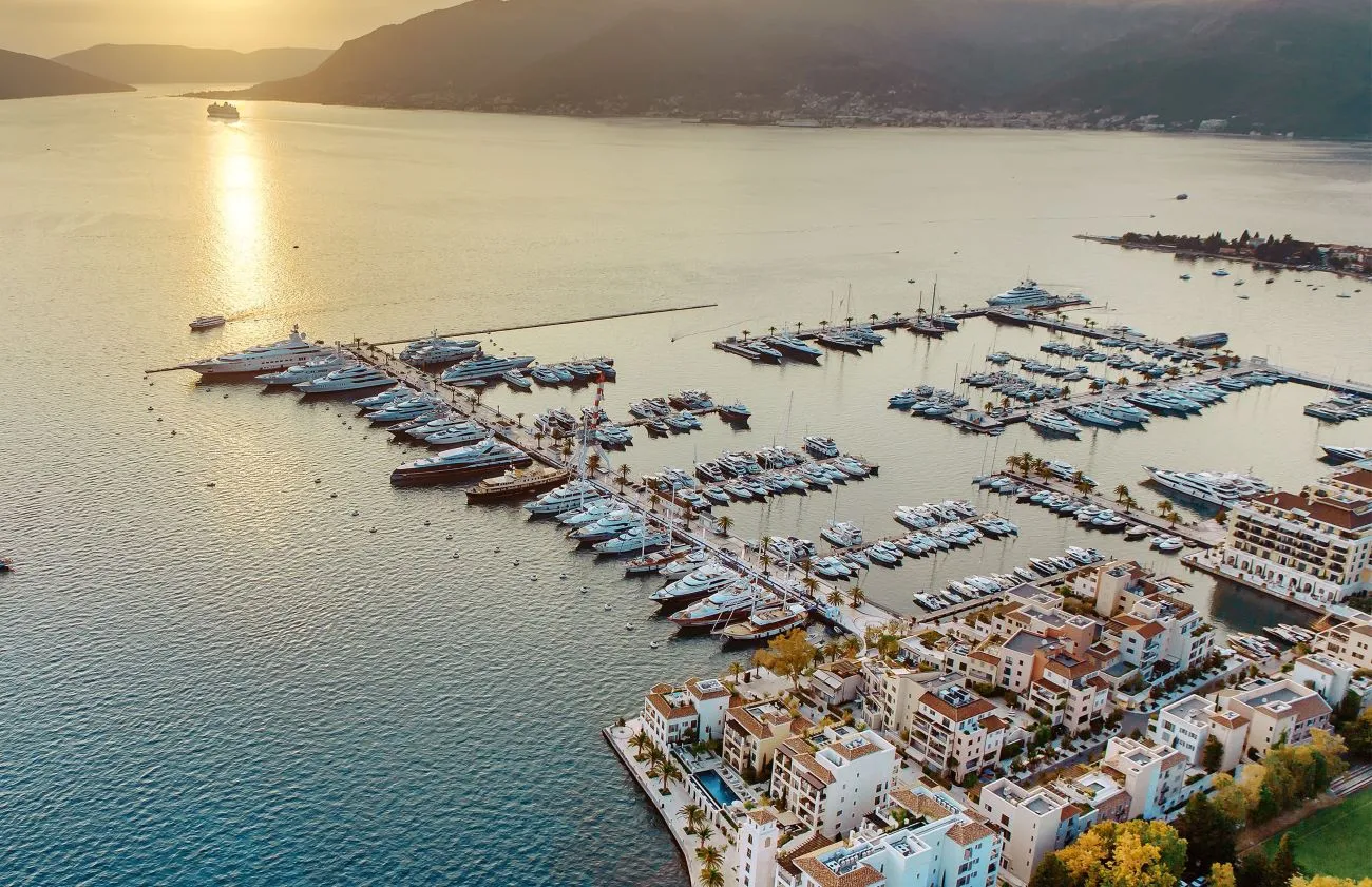 An aerial view of Porto Montenegro Marina, filled with superyachts and surrounded by luxury waterfront residences, with the Adriatic Sea stretching into the horizon under a golden sunset.