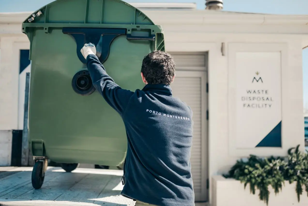 A worker wearing a navy blue "Porto Montenegro" fleece jacket is handling a large green waste bin outside a designated waste disposal facility. The facility has a white sign with the "M" logo, indicating an organized waste management system.