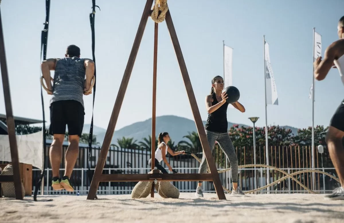 A group of people engaged in an intense outdoor workout session at Porto Montenegro Yacht Club, using suspension rings, medicine balls, and battle ropes against a scenic mountain backdrop.