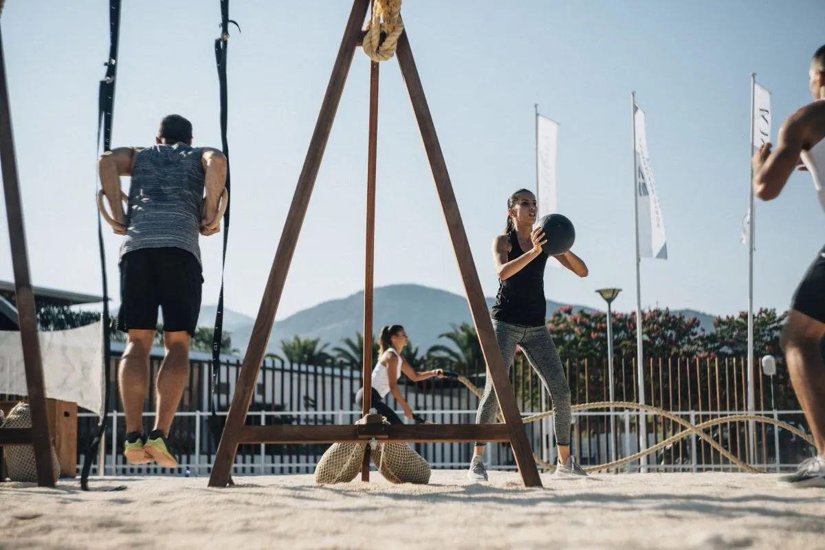 A group of people engaged in an intense outdoor workout session at Porto Montenegro Yacht Club, using suspension rings, medicine balls, and battle ropes against a scenic mountain backdrop.