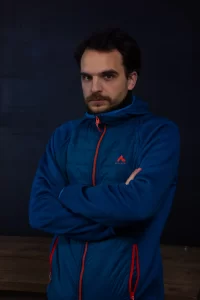 Aleksandar Ostojic posing with arms crossed, wearing a blue jacket with red accents against a dark background.