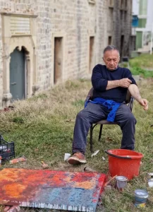 Portrait of Radun Marsenic seated outdoors, rolling up his sleeve next to a colorful painting and art supplies.