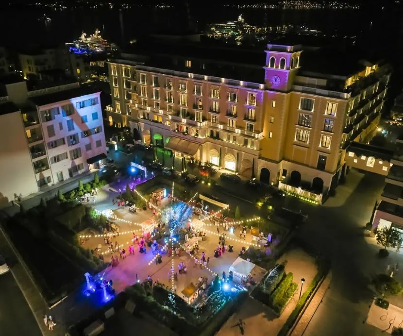 Aerial view of a lit-up Summer Garden of Reget hotel in Porto Montenegro, event with string lights, guests, and surrounding buildings.
